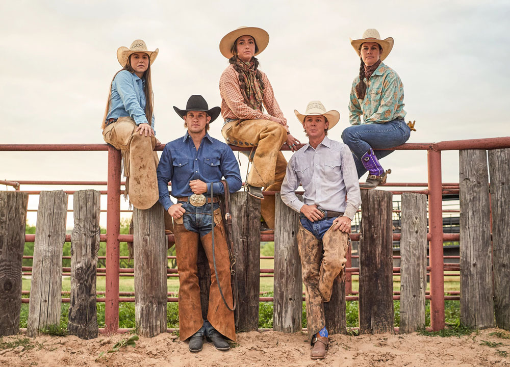 Tres mujeres y dos hombres en un rancho vestidos con jeans, camisa, sombrero de vaquero y botas de vaquero.